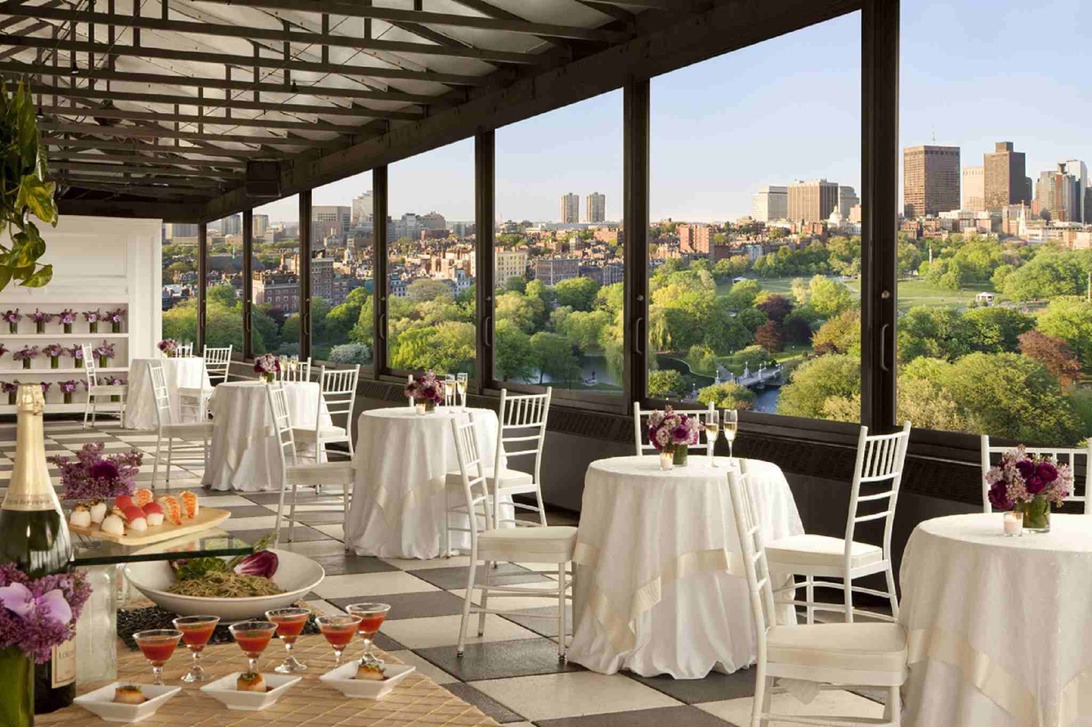 Elegant restauranglokal på hög höjd, förberedd för ett företagsevent med cocktailbord, vita dukar och lila blomsterarrangemang. Stora fönster erbjuder en vidsträckt utsikt över stad och park.