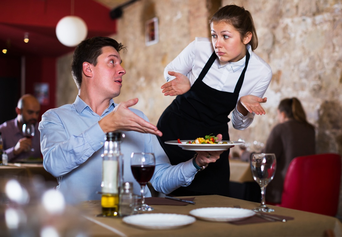 Servitör i professionell klädsel (vit skjorta och svart förkläde) interagerar med en gäst i affärsklädsel i en restaurangmiljö med tegelväggar och andra matgäster i bakgrunden.